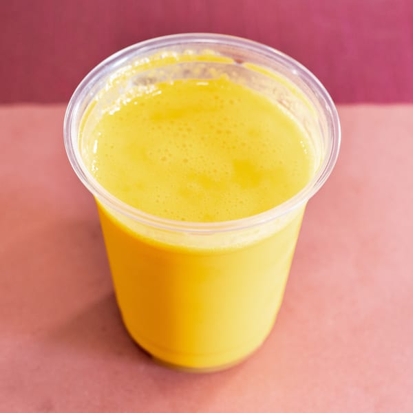 A vibrant yellow mango lassi in a clear plastic cup, set against a pink and maroon background.