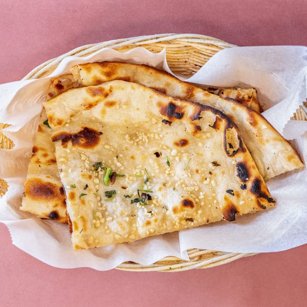 Basket of warm, golden-brown naan bread with garlic and herbs, served on a pink background.