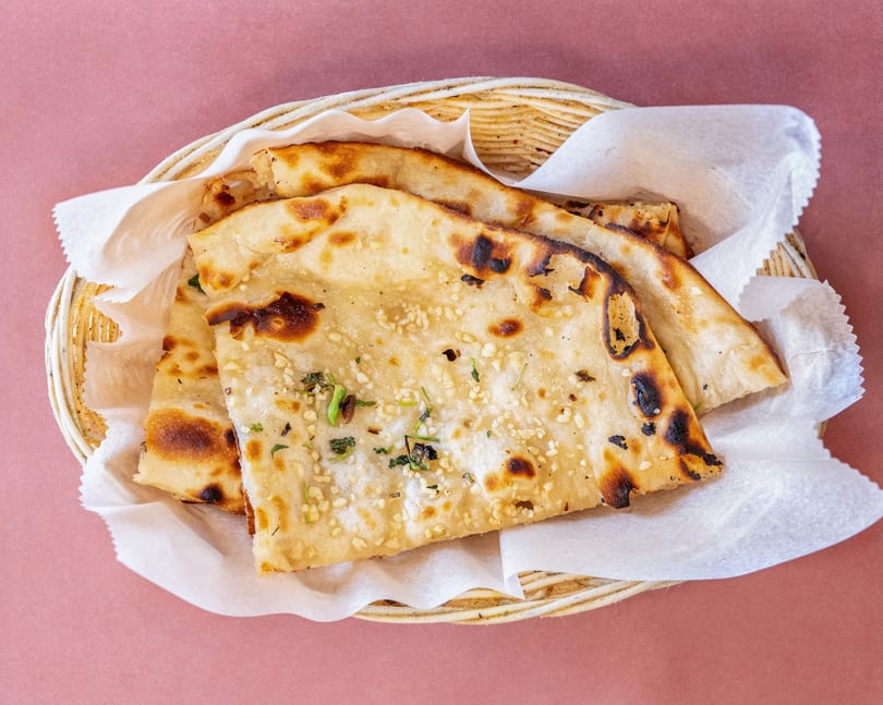 Basket of warm, golden-brown naan bread with garlic and herbs, served on a pink background.