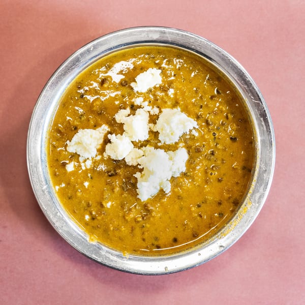 Overhead view of a metal bowl filled with yellow dal, topped with white paneer cheese.