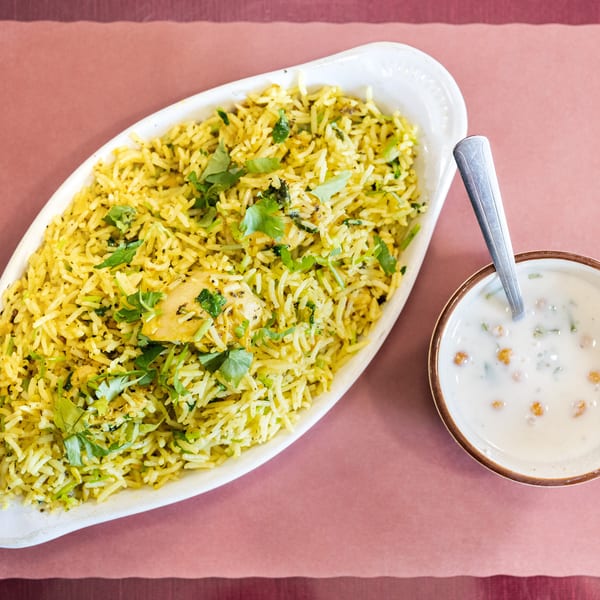 A long white dish of yellow rice with chicken and cilantro, next to a bowl of raita.