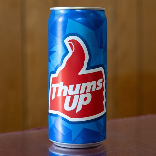 A blue Thums Up soda can with a red thumbs-up logo sits on a reflective brown surface.