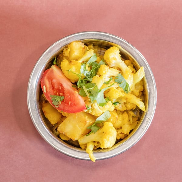 A metal bowl filled with yellow cauliflower and potato curry, garnished with cilantro and a tomato slice.