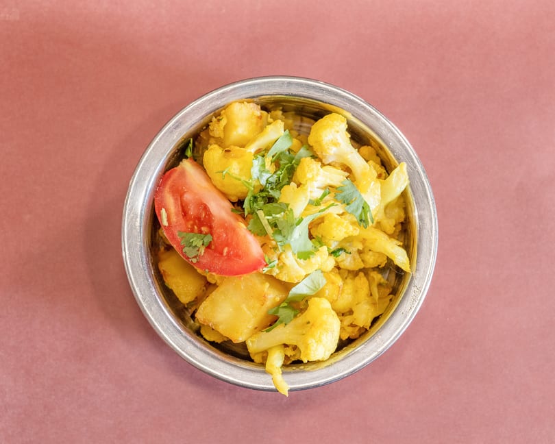 A metal bowl filled with yellow cauliflower and potato curry, garnished with cilantro and a tomato slice.